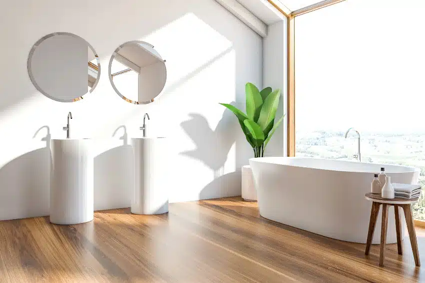 Scandinavian style bathroom with white walls, wooden floor, white bathtub standing near window and modern pedestal sinks with round mirrors