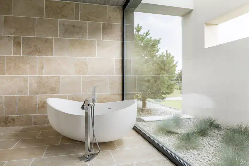 Spacious bathroom with bathtub, travertine tile wall, window, and freestanding faucet