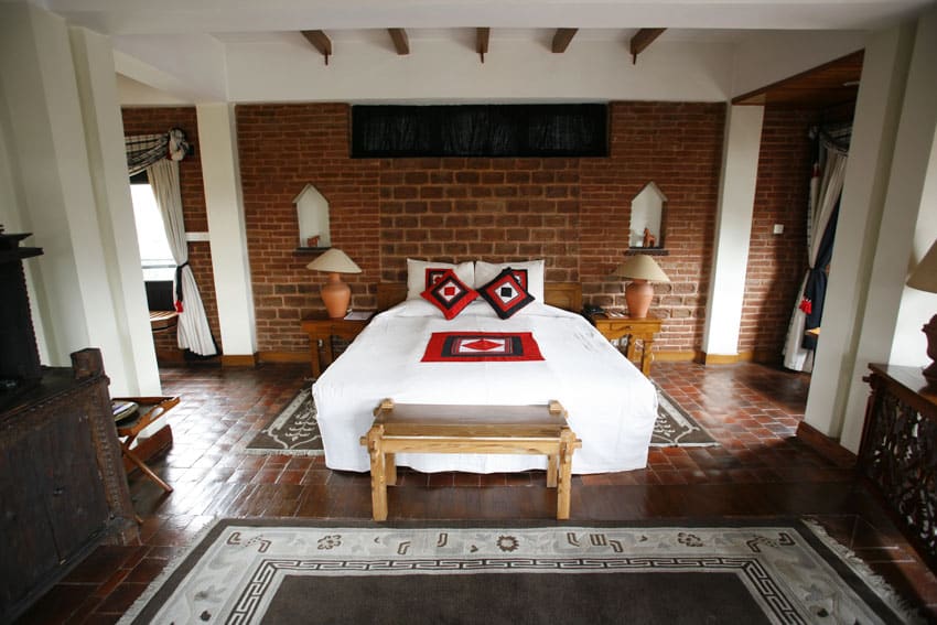 Spanish style bedroom with brick accent wall, nightstands, lamps, terracotta floor, rug, exposed ceiling beams, and dresser