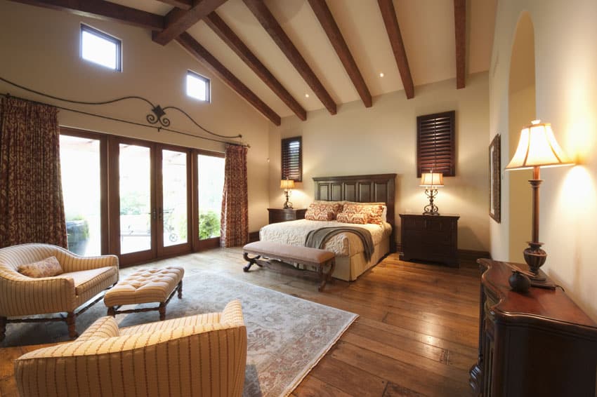 Spanish style bedroom with exposed ceiling beams, nightstands, lamps, wood flooring, dresser, chair, ottoman, wood floors, and glass door