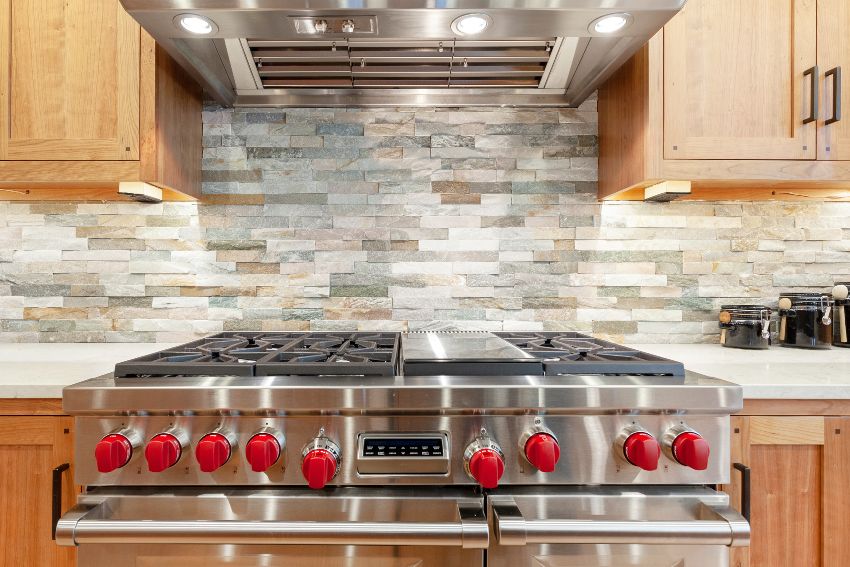 Stainless steel oven and stove with red knobs, stainless steel hood, brown kitchen cabinets and slate mosaic backsplash
