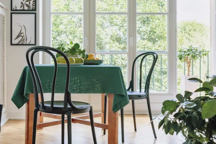 Two black chairs standing by dining table with apples, lemons, fresh plant and green tablecloth in bright room interior with two posters and balcony with view