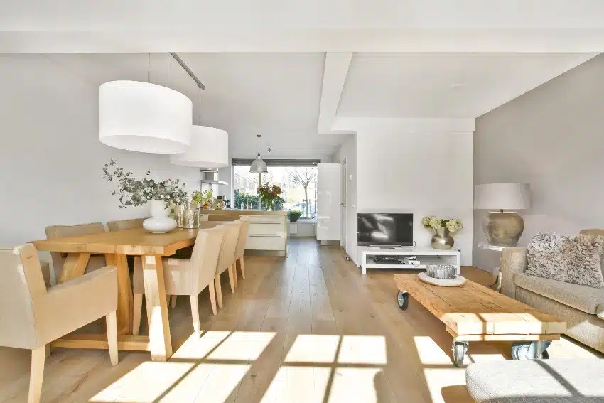 White and wooden interior of a home features a dining area with chestnut furniture, living area and a small kitchen in a modern house