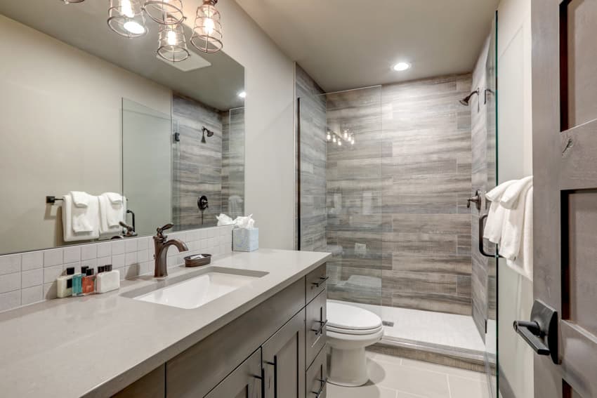 Bathroom with shower tile, glass divider, toilet, mirror, countertop, sink, faucet, and ceiling lights