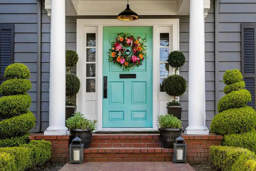 Beautifully decorated turquoise colored front door of traditional dark gray home with brick path and trimmed hedges