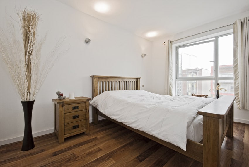 Bedroom with amish oak furniture, headboard, nightstand, floor vase, footboard, wooden flooring, ceiling lights, window, and curtains
