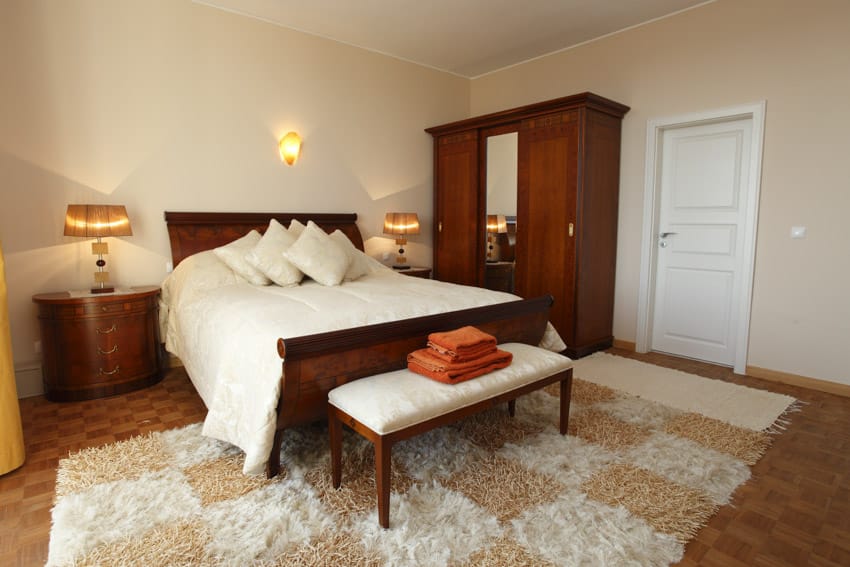 Bedroom with cherry wood nightstand, wardrobe, bench, carpet, bedding, headboard, wall sconce, and pillows