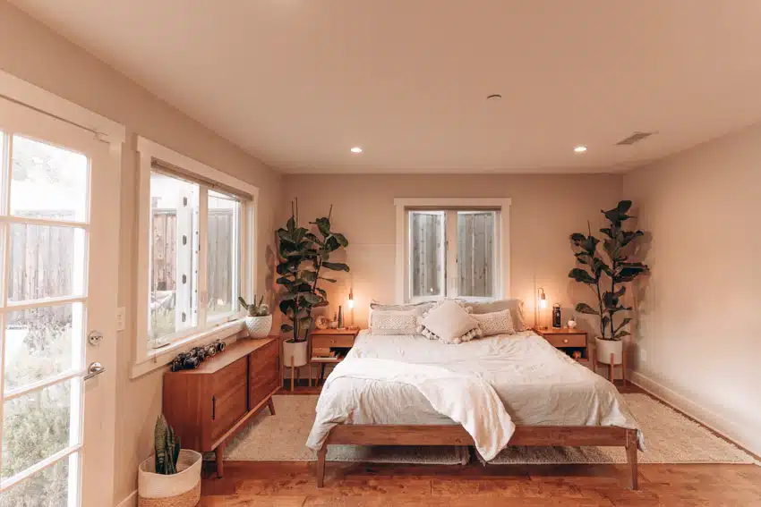 Bedroom with finished oak dresser, nightstands, bed frame, mattress, pillows, indoor plants, ceiling lights, and windows