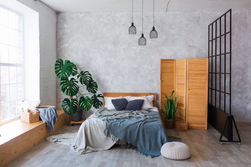 Boho bedroom with concrete wall, oak wood divider, bedding, ottoman, indoor plants, window, and pendant lights