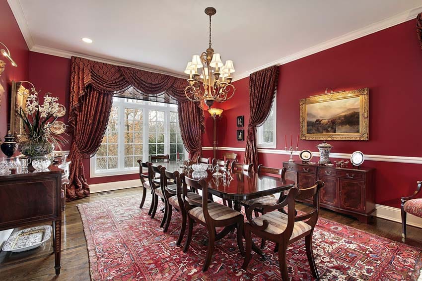 Classic dining room with red walls, cherry wood table, chairs, floor carpet, curtains, windows, buffet table, and chandelier