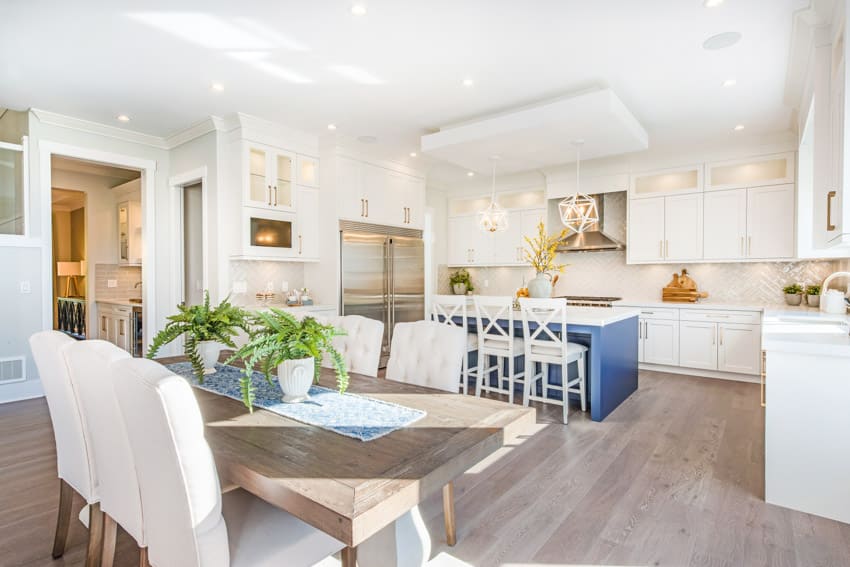 Coastal kitchen with wood dining table, chairs, island, countertop, white cabinets, backsplash, wooden flooring, and refrigerator