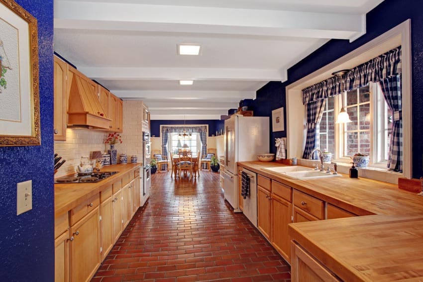 Farmhouse kitchen with wood countertop, cabinets, ceiling lights, windows, brick floors, and curtains