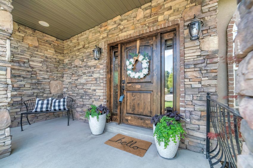 Front porch area with exterior ledger stone wall, wood door, wall sconces, bench, and potted plants
