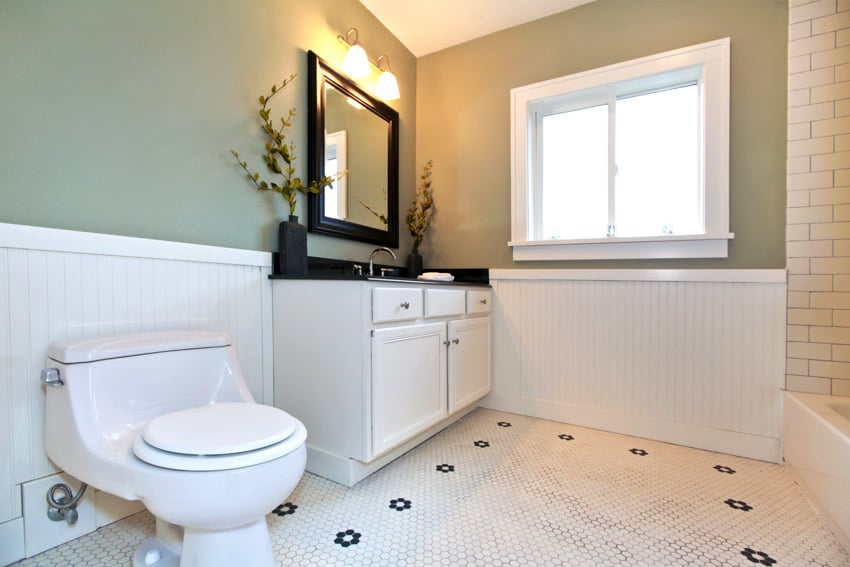 Green bathroom with toilet, tile floor, vanity mirror, black countertop, cabinets, accent lighting fixtures, and window