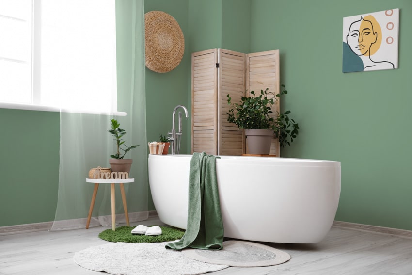 Green bathroom with tub, stool, laminate flooring, rug, room divider, indoor plant, window, and curtain