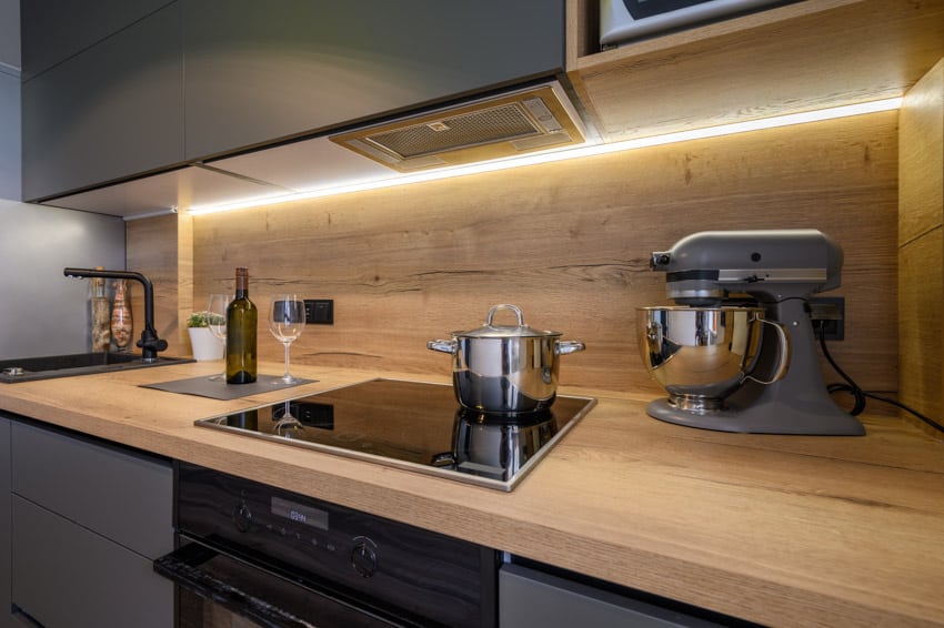 Modern kitchen with Indian teak countertop, gray cabinets, induction stove, wood backsplash, and under cabinet lighting