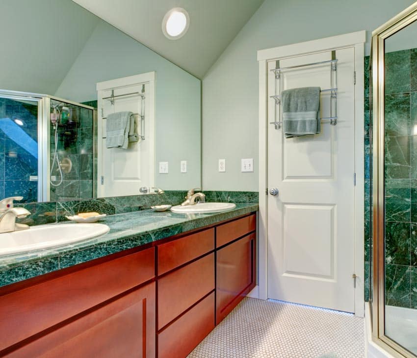 Seafoam green bathroom with sinks, mirror, wood cabinets, green marble countertop, and white door