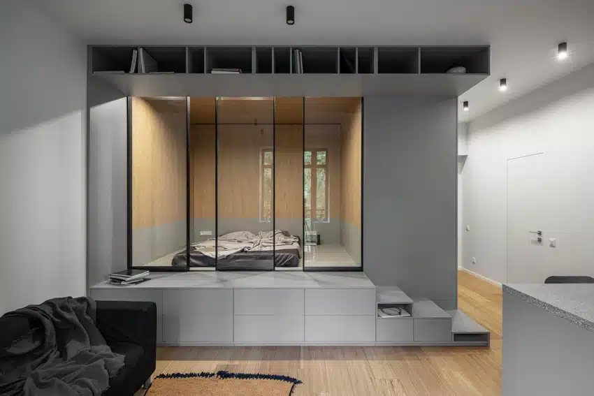 Small bedroom with bypass sliding doors, bed, mattress, wood walls next to living room with black couch