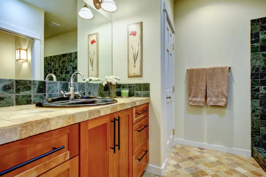 Soft green bathroom with wood cabinets, countertop, green tile backsplash, tile floors, sink, faucet, and mirror