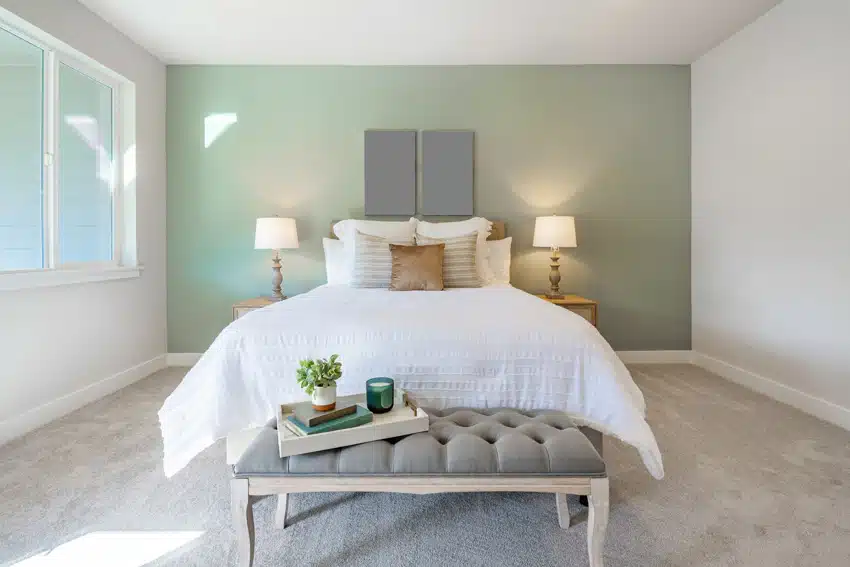Bedroom with green accent wall, nightstands, ottoman bench, lamps, comforter, pillows, and windows