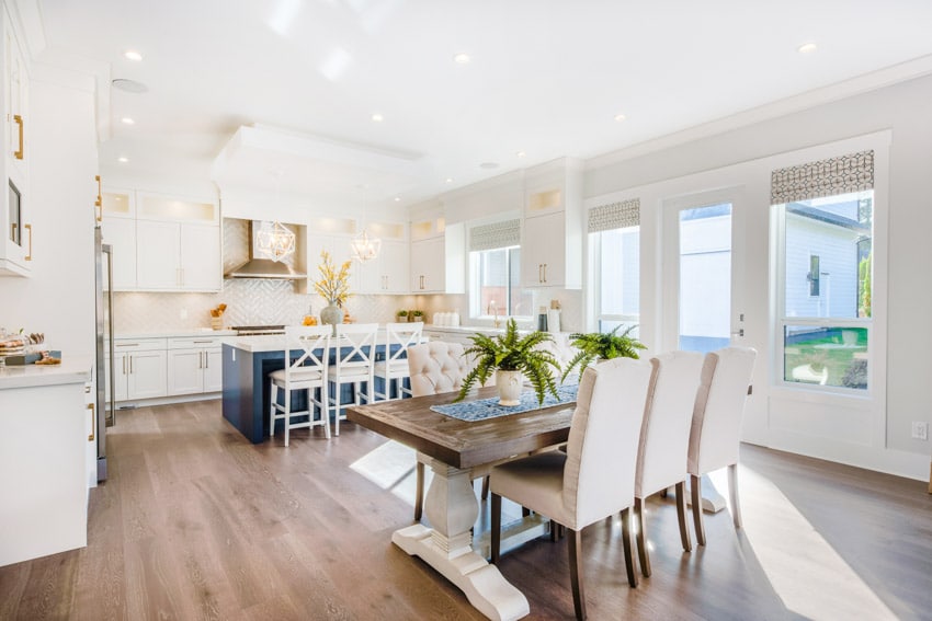 Coastal farmhouse kitchen with tile backsplash, wood flooring, dining table, chairs, island, high chairs, white cabinets, range hood, and glass windows