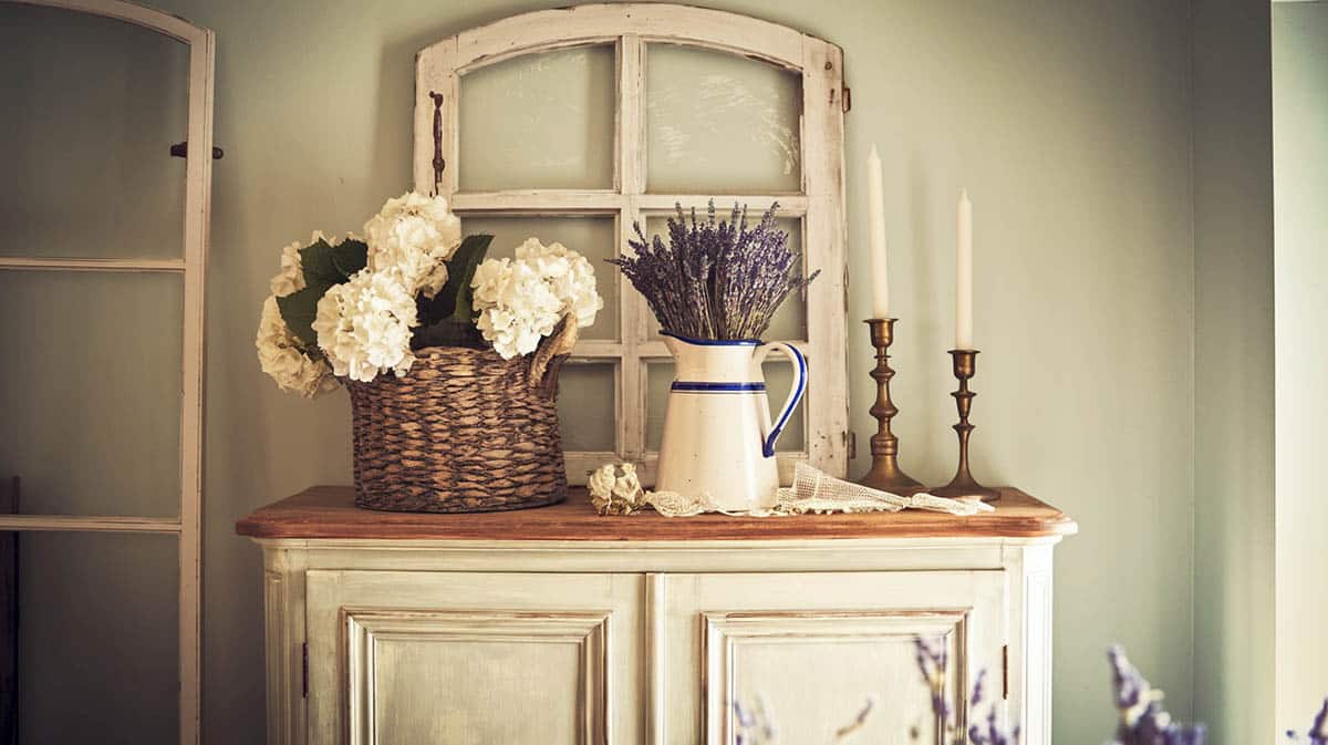 Weathered white french country armoire with enamelware with lavender brass candlesticks and wicker basket with flowers